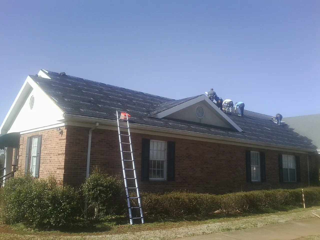 Crew tearing shingles off a brick ranch roof with a ladder set — Metal Roof Repair work by our crew in Potterville