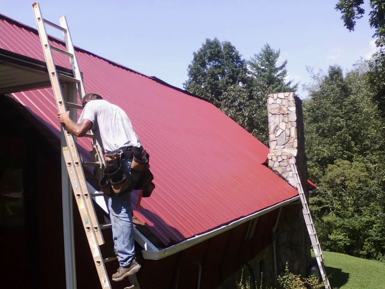 Roofer on a ladder beside a red metal roof and stone chimney — Soffit & Fascia Repair by Roofing Brothers in Potterville